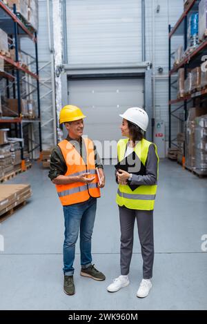 Employés d'entrepôt ayant une conversation amicale, homme et femme heureux dans des casques de sécurité et gilets de sécurité Banque D'Images