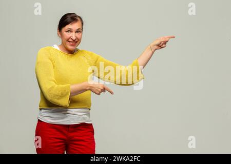 Une femme d'âge moyen pointe vers la droite avec sa main sur un fond gris. Banque D'Images