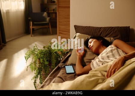 Jeune femme latino fatiguée en pyjama rayé faisant la sieste sur un lit confortable tout en étant allongée sous une couverture et en gardant la tête sur coussin Banque D'Images