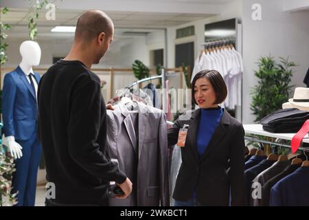 Boutique de mode client arabe demandant à l'assistante de femme asiatique pour opinion tout en choisissant la veste formelle. Vendeur et acheteur de magasin de vêtements tenant des vêtements sur cintre et discutant le style Banque D'Images