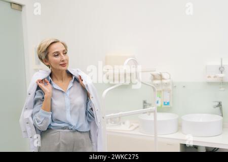 Un médecin féminin élégant d'âge moyen entre dans le bureau, retire la robe du cintre et le met. Le praticien professionnel se prépare à travailler en clinique Banque D'Images