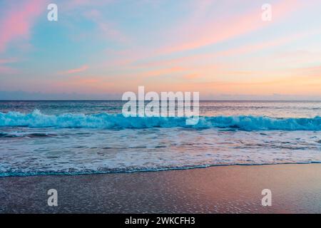 Le soleil se couche sur la plage tranquille, projetant des teintes chaudes à travers le ciel et reflétant sa lueur rose et dorée sur les vagues qui se détendent doucement Banque D'Images