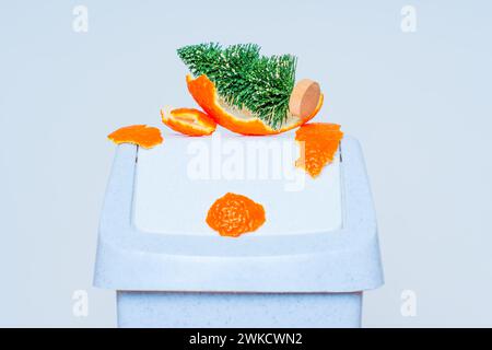 Figurine de sapin de Noël miniature mise au rebut repose parmi des pelures de mandarine sur une poubelle. Adieu aux Fêtes et au début du processus de nettoyage Banque D'Images