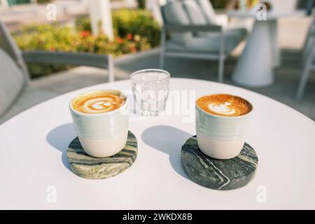 Deux tasses de délicieux café et un verre de glace sur une table blanche dans un café d'été par une journée ensoleillée Banque D'Images