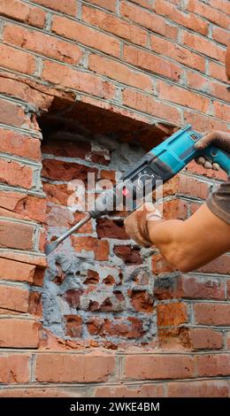 de fortes mains masculines ciselent la brique d'un mur de bâtiment avec une perceuse à percussion, photo verticale, brisant la surface de brique d'une maison à l'extérieur Banque D'Images