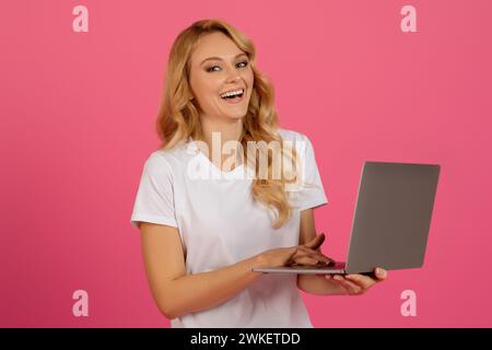 Femme blonde heureuse tenant un ordinateur portable sur fond de studio rose Banque D'Images