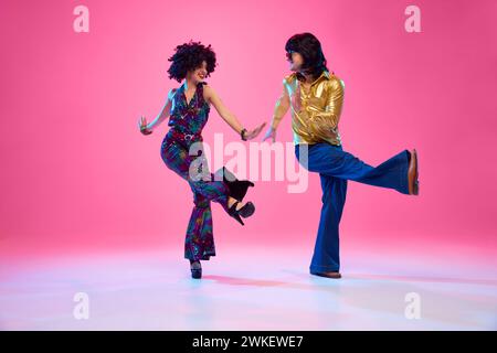 Homme et femme habillés dans une tenue vibrante des années 1970 dansant en mouvement sur fond de studio rose dégradé. Mode rétro. Banque D'Images