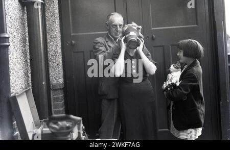 années 1940, historique, temps de guerre, une écolière, tenant un chat de compagnie, regarder sa grand-mère essayer un masque à gaz, Angleterre, Royaume-Uni. À la fin des années 1930, avec la guerre avec l'Allemagne à l'horizon, le gouvernement britannique a émis des masques à gaz pour le grand public, comme protection contre le largage de bombes à gaz positionnées, car les souvenirs étaient encore forts des horreurs des attaques au gaz qui ont dupé la première Guerre mondiale. Banque D'Images