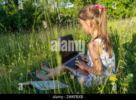 Une petite fille mignonne est assise dans un parc au milieu de fleurs et de types sur un ordinateur portable Banque D'Images