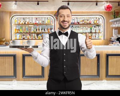 Portrait en longueur d'un serveur tenant un plateau avec des tasses à café et montrant les pouces vers le haut à un bar Banque D'Images