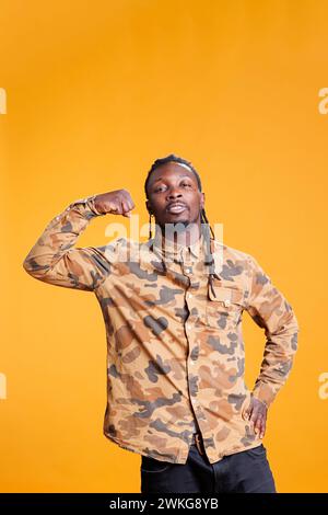 Portrait de bodybuilder athlétique montrant les muscles du bras entraînés devant la caméra en studio sur fond jaune. Homme afro-américain appréciant les exercices sportifs, s'entraînant à la force des triceps. Banque D'Images