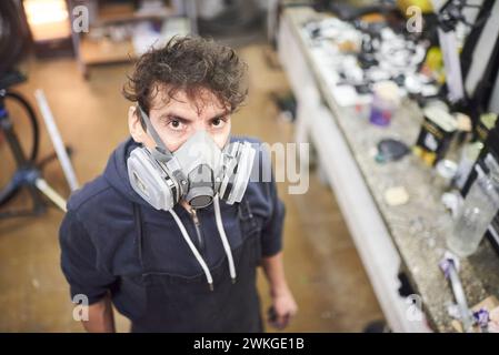 Portrait d'un jeune travailleur latin portant un masque de protection respiratoire regardant la caméra dans son atelier. De vraies personnes au travail. Composition de vue à grand angle Banque D'Images