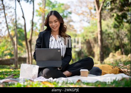 Une femme d'affaires asiatique positive et attrayante travaillant à distance dans un parc, travaillant sur sa tablette numérique sur un tapis de pique-nique. Banque D'Images