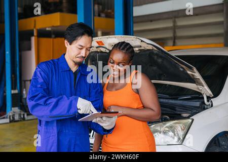 Les femmes clientes noires multiethniques heureuses apprécient avec la liste de contrôle de service de voiture de travailleur de mécanicien asiatique dans l'atelier de centre automatique Banque D'Images