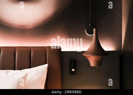 Cette image met en valeur une partie d'une chambre d'hôtel contemporaine avec une tête de lit moelleuse, une élégante suspension lumineuse et un décor mural artistique qui ajoute une touche Banque D'Images
