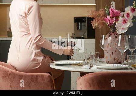 Femme en robe rose élégante tenant un verre à vin sur la table à manger dans l'appartement Banque D'Images