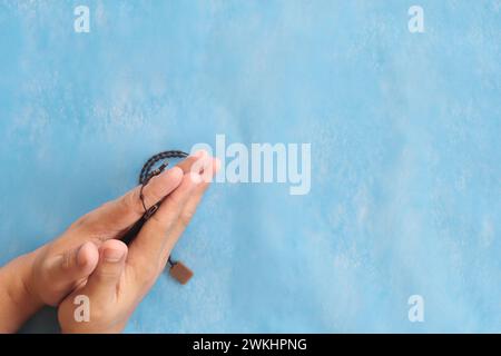 Mains masculines serrées ensemble, tenant et priant le chapelet ou le scapulaire sur fond bleu ciel. Banque D'Images