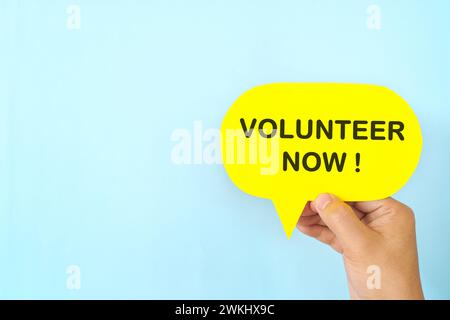 Main humaine tenant la signalisation jaune bulle de parole avec la phrase écrite Volunteer Now. Concept de volontariat. Banque D'Images