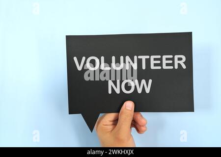 Main humaine tenant la signalisation jaune bulle de parole avec la phrase écrite Volunteer Now. Concept de volontariat. Banque D'Images