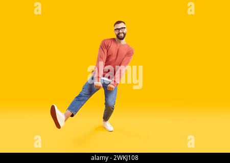 Portrait pleine longueur d'homme barbu drôle en sweat-shirt rouge et jeans dansant sur fond jaune. Banque D'Images