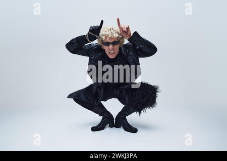 Jeune homme en tenue noire et bottes, gestuelle rock sur, avec une attitude vive et rebelle sur fond gris studio. Banque D'Images