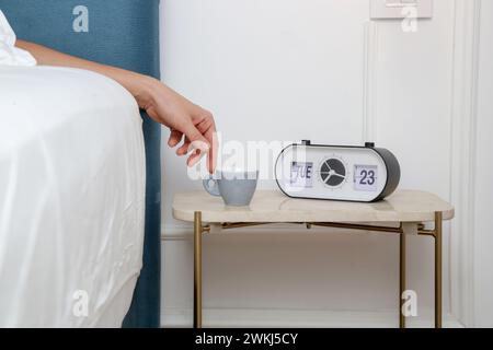 Femme au lit atteignant son premier café du matin de la table de nuit. Une tasse à espresso bleu clair sur la table de chevet à côté du réveil Banque D'Images