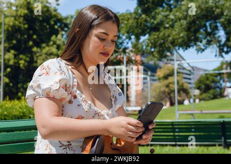 jeune femme violoniste buske latina assise sur un banc en bois à l'extérieur, vérifiant calmement les réseaux sociaux en utilisant le téléphone, la connexion des gens, la musique et la technologie Banque D'Images