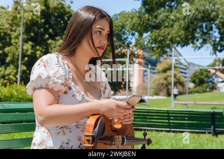 jeune femme violoniste buske latina assise sur un banc en bois à l'extérieur, vérifiant calmement les réseaux sociaux en utilisant le téléphone, la connexion des gens, la musique et la technologie Banque D'Images
