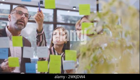 Équipe de gens d'affaires caucasiens debout près de la fenêtre en verre avec des notes collantes. Méthodologie Scrum avec des autocollants colorés. Collègues de travail discutant p Banque D'Images