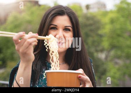 Jeune femme mangeant des nouilles avec des baguettes sur un fond urbain vert - concept de restauration rapide et décontracté - jeune femme appréciant la cuisine asiatique Banque D'Images