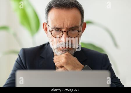 Un homme mûr réfléchi dans un costume décontracté élégant avec des lunettes, se concentrant profondément sur son ordinateur portable Banque D'Images