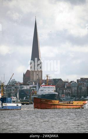 21 février 2024, Mecklembourg-Poméranie occidentale, Rostock : L'épave du navire de la station balnéaire 'Undine' est remorquée le long de la Warnow en direction du port de pêche de Marienehe par des remorqueurs, avec tous L'église de Pierre en arrière-plan. Le navire, qui a été amarré dans le port de la ville pendant dix ans, est considéré comme le plus ancien navire de station balnéaire en Allemagne. Il a été construit en 1910. Le 'Undine' doit être remorqué du port de la ville au port de pêche. Il sera ensuite sorti de l'eau avec une grue et mis à terre. Selon les estimations de Rostock, la restauration de la barge de longue durée Banque D'Images