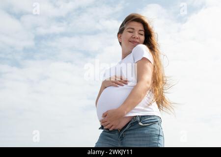Heureuse femme enceinte caucasienne tenant son ventre sur le fond du ciel. t-shirt blanc et jeans. Troisième trimestre. Concept de grossesse heureuse Banque D'Images