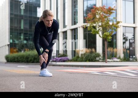 Femme athlétique dans la douleur tenant son tibia, montrant des signes de blessure tout en faisant du jogging dans un environnement urbain. Banque D'Images