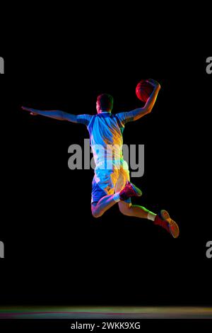 Portrait dynamique d'un joueur de basket-ball faisant une superposition parfaite en action sur fond noir de studio dans une lumière néon mixte. Banque D'Images