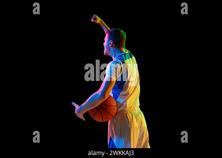 Portrait de vue arrière d'un jeune sportif tient une balle de basket-ball et crie sur fond noir de studio dans une lumière de néon mixte. Banque D'Images