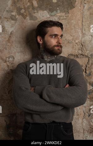 beau mannequin homme brutal avec une barbe et une coiffure dans un pull tricoté vintage à la mode se tient près d'un mur de béton grunge Banque D'Images