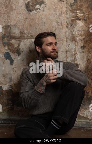 beau jeune homme hipster brutal avec une coiffure et une barbe avec un tatouage dans un pull tricoté de mode et jeans noir se trouve près d'un vieux concre grunge Banque D'Images