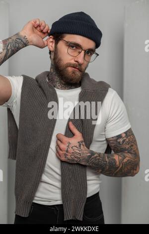 Cool élégant jeune homme hipster avec une barbe et des tatouages avec des lunettes de mode dans un T-shirt blanc avec un pull met sur un chapeau tricoté sur un dos blanc Banque D'Images