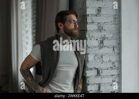 Bel homme hipster réussi avec tatouage et barbe dans des vêtements de mode avec un T-shirt blanc et un pull se tient près de la fenêtre dans la pièce Banque D'Images