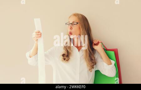 Choqué et surpris femme d'âge moyen caucasien dans des lunettes semble étonné par l'étiquette de prix élevé sur le long reçu papier tenant sac à provisions Banque D'Images