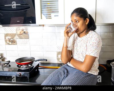 Une jeune femme sud-asiatique en pyjama confortable sourit brillamment en commençant sa journée avec un verre d'eau Banque D'Images