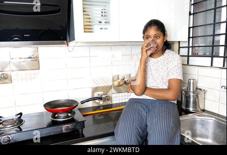 Une jeune femme sud-asiatique en pyjama confortable sourit brillamment en commençant sa journée avec un verre d'eau dans une cuisine élégante Banque D'Images