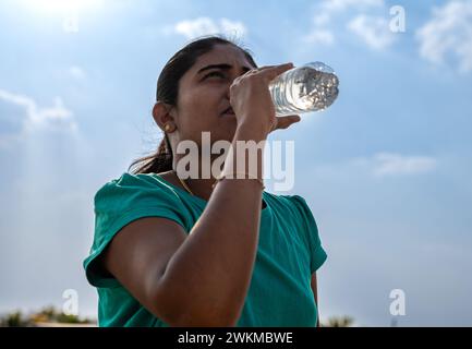 Une jeune femme reprend son souffle et se réhydrate avec de l'eau après un jogging rafraîchissant dans un parc animé Banque D'Images