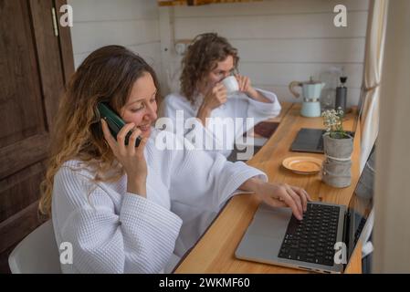Une femme dans un peignoir blanc sourit tout en parlant au téléphone et en travaillant sur son ordinateur portable dans un bureau à domicile, avec une autre personne en arrière-plan sirotant un verre. Banque D'Images