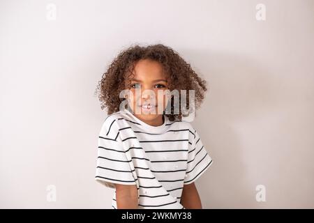 Une fille afro-américaine aux cheveux bouclés dans une chemise rayée sourit joyeusement et regarde la caméra, sur un fond blanc simple, heureuse enfance A. Banque D'Images