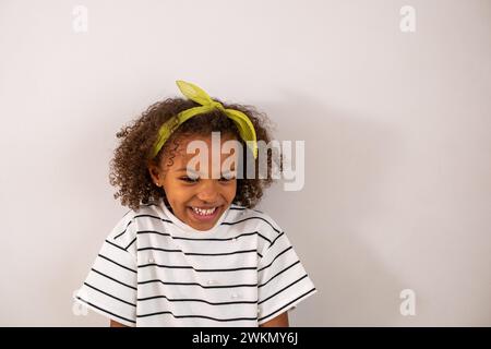 Une fille afro-américaine aux cheveux bouclés dans une chemise rayée et un bandeau citron rit joyeusement, sur un fond blanc simple, enfance heureuse. Haute qualité Banque D'Images
