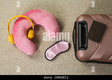 Oreiller de voyage rose avec écouteurs, valise, passeport et masque de sommeil sur tapis beige, plat Banque D'Images