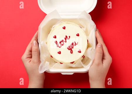 Femme tenant une boîte à emporter avec un gâteau au bento à la table rouge, gros plan. Prog Surprise pour la Saint-Valentin Banque D'Images