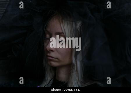 Le visage d'une jeune femme blonde caucasienne est partiellement obscurci par un voile sombre, les yeux fermés dans une expression sereine Banque D'Images